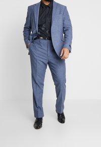 Homme en costume bleu avec une chemise noire à motif floral, les mains dans les poches, debout devant un fond blanc uni.