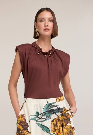 Frau mit kurzen braunen Haaren trägt ein bordeauxfarbenes ärmelloses Oberteil mit Perlendetails und weiße weit geschnittene Hose mit Blumenmuster, Hände in den Taschen.