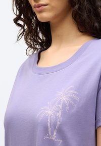 Γυναίκα που φοράει μωβ πουκάμισο με γραφικό φοίνικα και το κείμενο "slow living" στο στήθος, με  σγουρά σκούρα μαλλιά και ουδέτερη έκφραση.