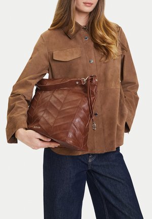 Žena v hnědém semišovém kabátu a tmavých džínách drží velkou hnědou koženou kabelku se vzorem chevron a kovovým přívěskem.