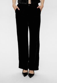 Schwarze Samt-Hose mit weitem Bein, glatter Textur, doppelten Seitentaschen und Knopfverschluss, kombiniert mit schwarzen spitzen Pumps.