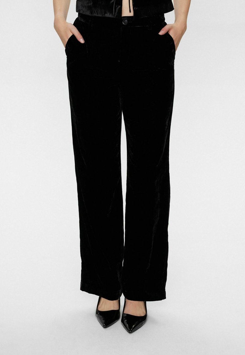 Schwarze Samt-Hose mit weitem Bein, glatter Textur, doppelten Seitentaschen und Knopfverschluss, kombiniert mit schwarzen spitzen Pumps.