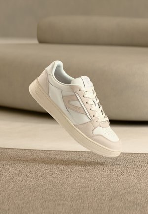 Λευκό και μπεζ sneaker χαμηλού προφίλ με κορδόνια, ακουμπισμένο πάνω σε μπεζ χαλί και μινιμαλιστικό μπεζ καναπέ στο φόντο.