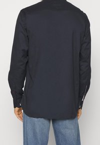 Granatowa koszula z długim rękawem, z zapinanymi mankietami, gładką fakturą, spiczastym kołnierzykiem oraz subtelnym czerwono-białym akcentem w pobliżu mankietów. Zobacz widok z tyłu.