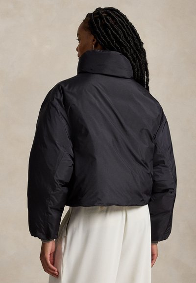 Personne aux longs cheveux tressés, portant une doudoune noire et un pantalon blanc, vue de dos sur un fond uni.