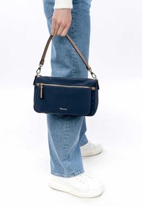 Marineblaue Umhängetasche aus Stoff mit einer Vorderreißverschlusstasche, braunem Lederhenkel und rechteckiger Form. Glatte Textur, minimale Details.