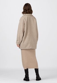 Beige ullblandad oversized kappa med krage, i kombination med en ribbad beige midikjol och svarta ankelboots, visad bakifrån.