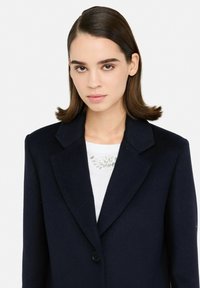 Blazer di lana blu navy con colletto a rever e chiusura con un solo bottone. Indossato sopra una maglietta bianca con decorazioni in cristallo.
