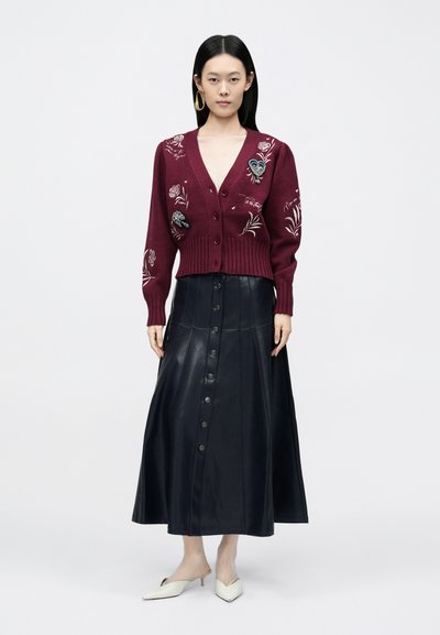 Bordowy kardigan z kwiatową haftą, dekoltem w serek i guzikami, w połączeniu z czarną skórzaną długą spódnicą zapinaną na guziki z przodu. Białe spiczaste baleriny.