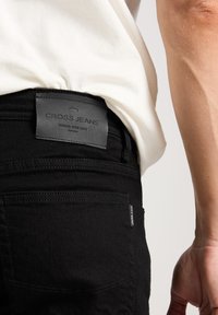 Zwarte denim jeans met een leren label met de tekst "CROSS JEANS" bevestigd aan de tailleband. Kenmerken zijn onder andere achterzakken en subtiele stiksels.