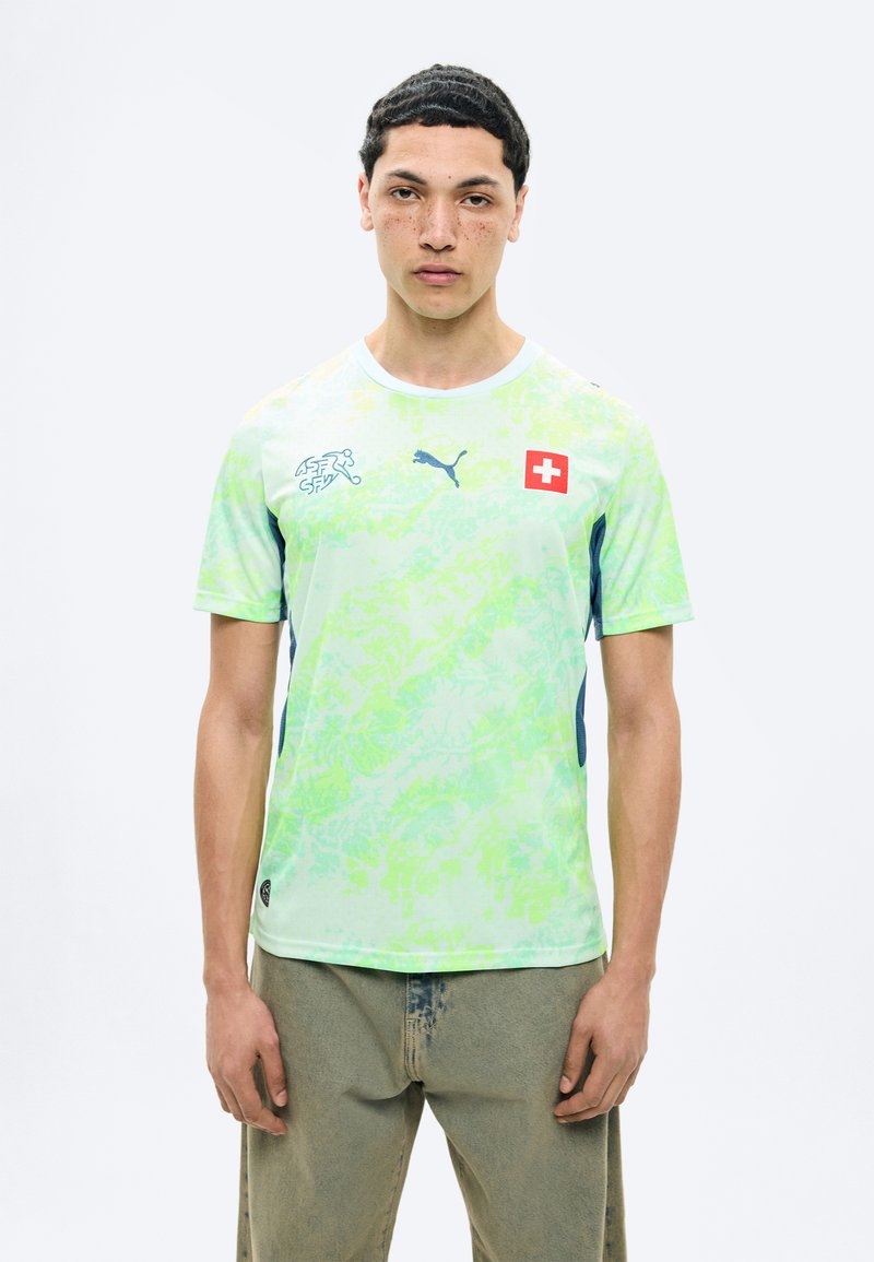 Junger Mann trägt ein hellgrünes, gemustertes Sporttrikot mit Schweizer Flagge, Puma-Logo und beigen Hosen, steht vor einem schlichten weißen Hintergrund.
