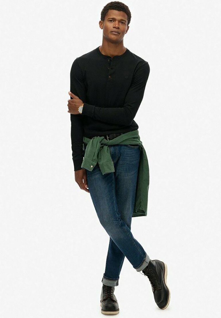 Zwarte langharige henley-shirt, donkerblauwe jeans, groene shirt geknoopt om de taille, en zwarte leren laarzen met een tan/kleurige zool.