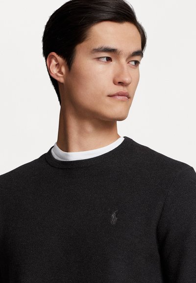 Νεαρός άνδρας με σκούρα μαλλιά, φορώντας ένα μαύρο υφασμάτινο πουλόβερ πάνω από ένα λευκό πουκάμισο, κοιτάζοντας προς τα δεξιά σε μια απλή φόντο.