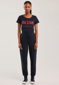 Moteris stovinti vilkinti juodą marškinėlį su užrašu „BIG STAR Your Life. Your Jeans.“, juodus raištelius turinčius bėgimo kelnes ir juodus aukštakulnius kedus.