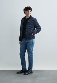 Navy quilted jacket with a stand collar, paired with a black sweater and blue jeans. Model wears black leather shoes, standing against a neutral background.