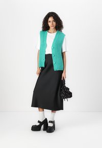 Turquoise knitted vest with buttons over a white T-shirt, paired with a long black skirt and chunky black platform shoes.