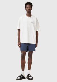 Yli suurikokoinen valkoinen puuvillapaita mustalla logolla, yhdistettynä laivastonsinisiin shortsihousuihin. Mallilla on päällään mustat sandaalit kiiltävin yksityiskohdin ja valkoiset sukat.