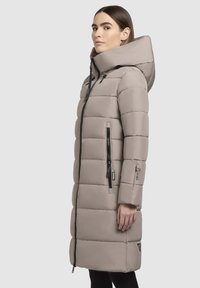 Long puffer coat in light beige, featuring a hood, front zipper, and side pockets with black zippers. Quilted design with smooth texture.
