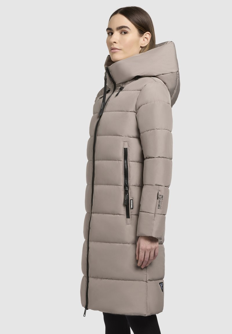 Langer Puffer-Mantel in hellbeige, mit Kapuze, Front-Reißverschluss und Seitentaschen mit schwarzen Reißverschlüssen. Gequiltes Design mit glatter Textur.