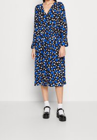 Abito con motivo blu e bianco su sfondo nero, con scollatura a V, maniche lunghe e vita elastica, abbinato a scarpe con plateau nere.