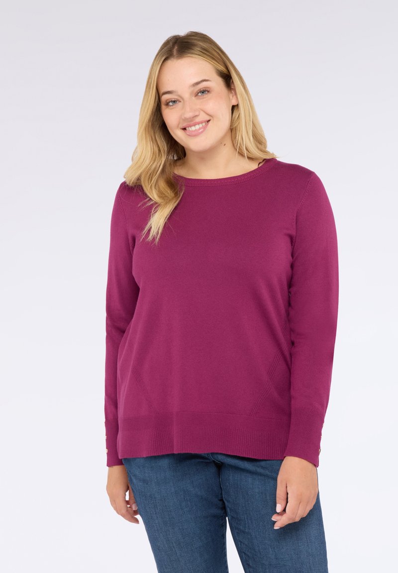 Mujer sonriente con cabello largo rubio, vistiendo un suéter de manga larga púrpura y jeans azules, frente a un fondo claro y liso.