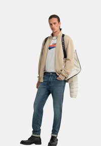 Giacca bomber beige sopra una maglietta bianca con grafica e strisce blu e rosse, jeans blu e stivali neri con lacci; una borsa trapuntata è appesa a una spalla.