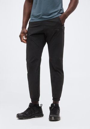 Személy fekete szűk szárú nadrágot és fekete sportcipőt visel, egyik kezét a zsebében tartva, egyszínű háttér előtt áll.