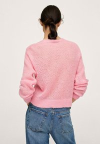 Pull en tricot rose avec un design ample et aéré et un ourlet côtelé. Présente des manches larges et un motif texturé, associé à un jean en denim bleu.