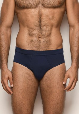 Torso masculino y piernas superiores vistiendo bóxers oscuros lisos, de pie contra un fondo neutro con los brazos relajados a los lados.