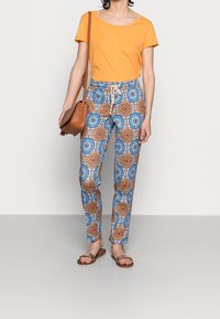 Orange t-shirt med kort ärm, mönstrade blå och bruna byxor med snörning, samt en brun axelväska och platta sandaler.