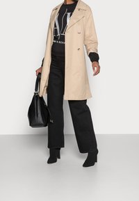 Trench beige sopra una felpa nera con stampa grafica, abbinata a pantaloni neri a gamba larga e stivaletti con tacco. Borsa nera con tracolla lunga.