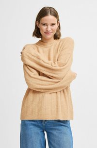 Beige pletený svetr s žebrovanou texturou, dlouhými širokými rukávy a klasickým kulatým výstřihem, doplněný o světlé modré džíny.