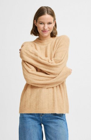 Pull en tricot beige avec une texture côtelée, doté de manches longues et larges ainsi qu'un col rond, associé à un jean bleu clair.