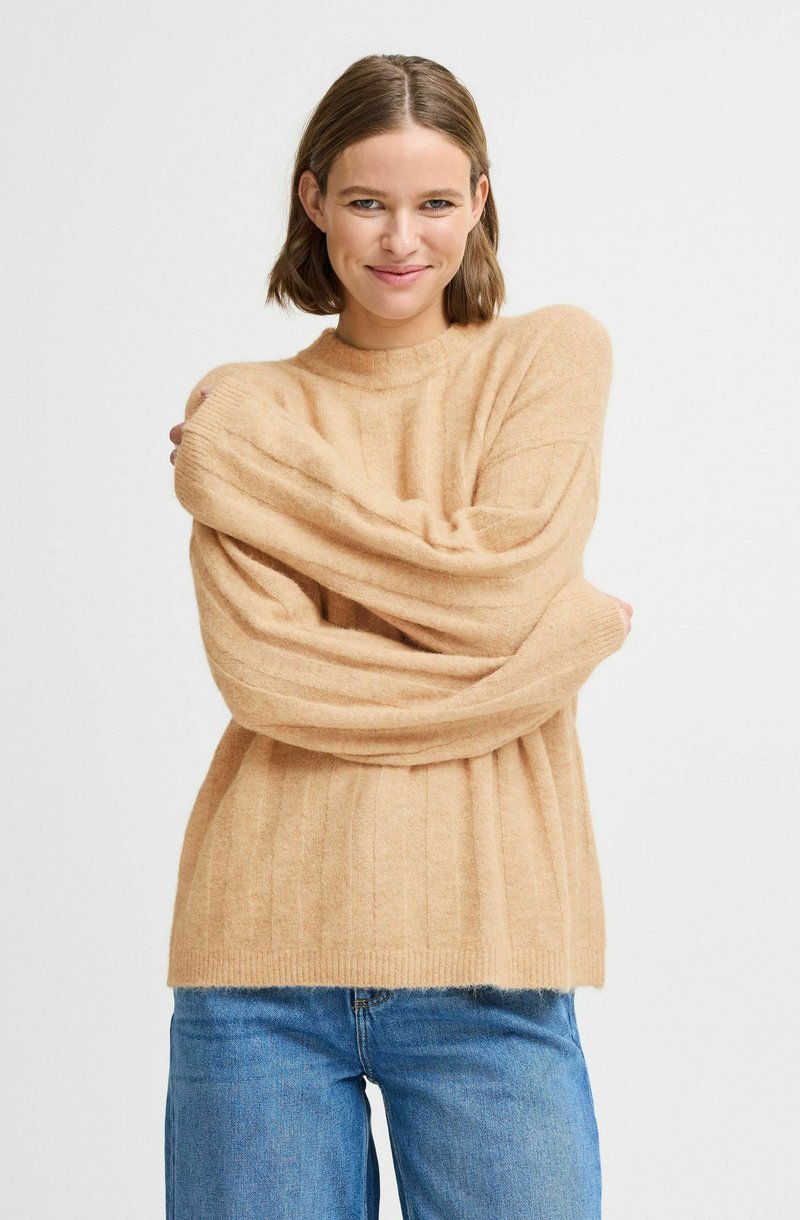 Beige stickad tröja med ribbat mönster, med långa, breda ärmar och rundad halsringning, kombinerad med ljusblå jeans.