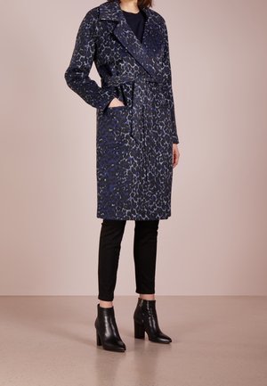 Femme portant un manteau léopard bleu foncé ceinturé, arrivant au genou, avec un pantalon noir et des bottines à talons noires.