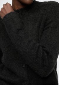 Czarny kardigan o miękkiej, puszystej fakturze, z okrągłym dekoltem, zapinany na guziki z przodu i długimi rękawami, widoczny częściowo rękaw.