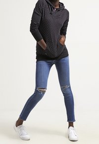 Sudadera negra con puntos blancos, bolsillo tipo canguro y capucha. Lleva puesta con jeans azules desgastados y zapatillas blancas.