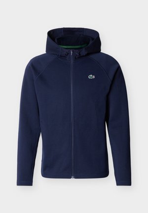 Tumesinine lukuga dressipluus kapuutsiga, millel on nööriga kapuuts ja vasakul rinnal väike roheline krokodillilogoga, esitatud valgel taustal.