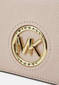 Szczegół jasnoróżowej skórzanej torebki z metalicznym złotym logo z inicjałami "MK" oraz wytłoczonym napisem "MICHAEL KORS" dookoła.