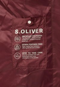 Bordeauxkleurige jas met een zak waarin witte tekst de materialen, onderhoudsinstructies en type isolatie beschrijft, gemaakt van gerecycled polyester.