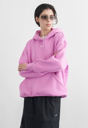Person trägt übergroßen rosa Hoodie und schwarzen Rock, mit kurzem dunklem Haar und Sonnenbrille, steht mit verschränkten Armen vor einfarbigem Hintergrund.