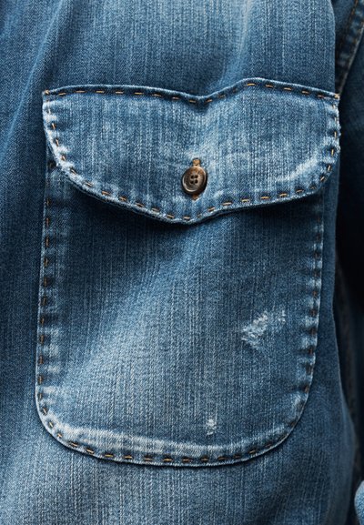 Μπλε τζιν τσέπη πουκάμισου με καπάκι που ασφαλίζει με καφέ κουμπί και εμφανείς ραφές σε ύφασμα με ελαφρώς ξεθωριασμένες και φθαρμένες λεπτομέρειες.