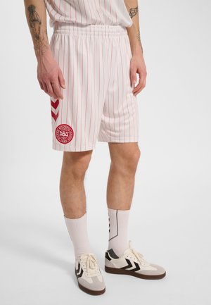 Mann trägt weiße Sportshorts mit roten Nadelstreifen und einem roten rundem Logo am linken Bein, kombiniert mit weißen Sportsocken und weißen Turnschuhen.