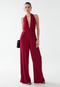 Mono rojo tipo halter con escote en V profundo y pantalones de pierna ancha. Tela lisa, detalle de pliegue frontal y combinado con un bolso negro de mano.