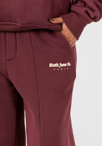 Bordeauxrode sweatpants met een zachte textuur, voorzien van zijzakken en een logo in lichtroze, geplaatst op de heup.