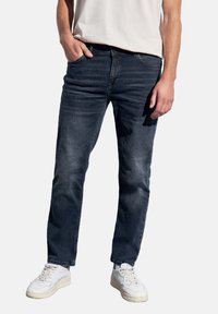 Hombre vistiendo jeans azul oscuro, zapatillas blancas y una camiseta blanca, con una mano en el bolsillo, de pie frente a un fondo simple.