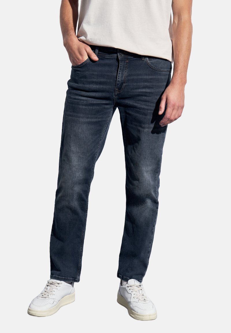 Hombre vistiendo jeans azul oscuro, zapatillas blancas y una camiseta blanca, con una mano en el bolsillo, de pie frente a un fondo simple.