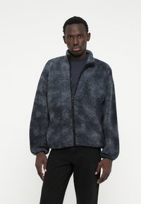 Dunkelgraue Fleecejacke mit strukturierter Oberfläche, hohem Kragen, durchgehendem Reißverschluss und elastischen Bündchen, kombiniert mit einem navyfarbenen Hemd und schwarzen Hosen.