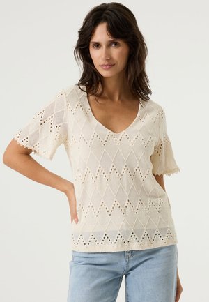 Vrouw met donker haar die een crèmekleurige, kortarmige gebreide top met diamanten patronen en lichtblauwe spijkerbroek draagt, staand tegen een effen achtergrond.