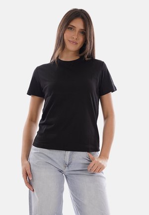 Giovane donna con capelli castani lisci che indossa una maglietta nera a maniche corte e jeans azzurri, in piedi con una mano in tasca.